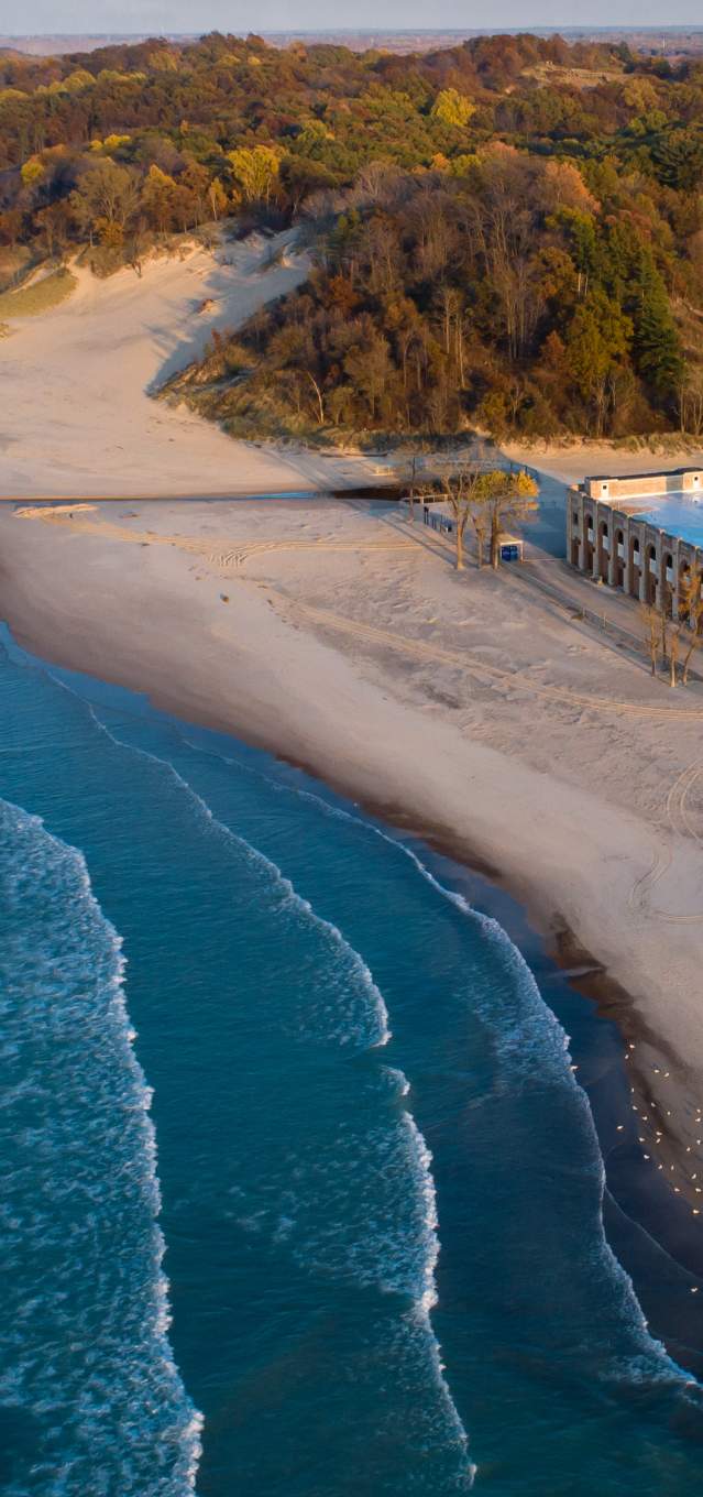 Bright blue water with white cresting waves near the shore sits to the left. On the right is a long sandy beach that spreads to the horizon line. Wooded dunes extend from the beach and into the distance. A stone building sits on the beach with a parking lot behind it.