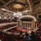 Inside an amphitheater from the balcony. White lights frame the stage and line the edges of the balconies. A small orchestra is playing to an audience