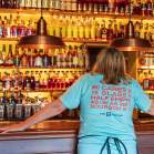 A woman in a light blue shirt stands in front of seemingly hundrands of bourbon botttles. Her shirt reads, "Who cares if the glass is half empty as long as there's bourbon in it" The B-Line