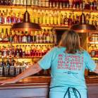 A woman in a light blue shirt stands in front of seemingly hundrands of bourbon botttles. Her shirt reads, "Who cares if the glass is half empty as long as there's bourbon in it" The B-Line