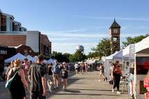People walk down the street lined with pop-up tent booths selling art with the Overland Park water tower and clock tower in the background