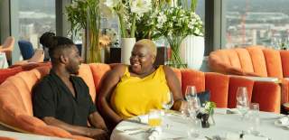 A man and woman sit talking at a table in a fancy restaurant, the window behind them looks out over Birmingham