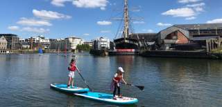 Children stand up paddle boarding by the SS Great Britain on the Bristol Harbourside