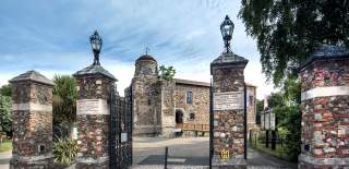 Colchester Castle photographed from outside the Castle Park gates. The Gates are open and four chunky stone pillars with lamps on top sit in the foreground.