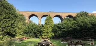 Wooden Tables sit on a lawn in a pub beer garden surrounded by greenery. A large, picturesque brick viaduct sits behind them in the middle distance