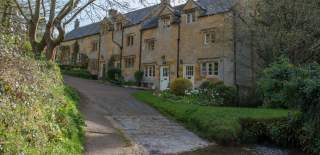 A row of traditional Cotswold stone cottages beside a narrow lane, with a shallow stream running alongside and trees and greenery in the foreground.