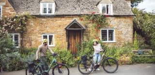 A couple push their bikes past a beautiful old cottage in a Cotswold village