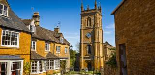 Narrow village street lined with honey-coloured stone cottages leading to the tall church tower of St Peter & St Paul church in Blockley, under a bright blue sky.