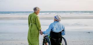 An elderly couple holds hands on a sandy beach. The woman in a green dress walks beside a man in a wheelchair, both gazing at the calm sea.