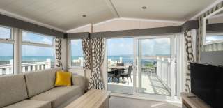 Bright living room with a beige sofa and yellow cushion, facing sliding glass doors that open to a balcony with a sea view. Airy and serene atmosphere.