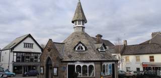 Grey stone building with a steep slate roof and spire, situated in a quaint village setting. Surrounding buildings are charming, evoking a peaceful, rustic feel.
