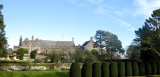 Historic stone manor with gabled roof set against a clear blue sky. Trimmed hedges and lush green lawns in the foreground create a serene atmosphere.