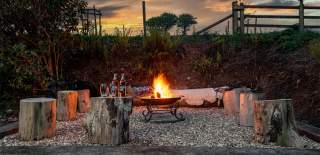 A cosy outdoor setting with a central fire pit surrounded by tree stump stools. Two beer bottles and glasses sit nearby. The sunset sky adds warmth.