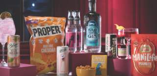 A selection of snacks and drinks is displayed against a red curtain backdrop. Items include popcorn, gin, peanuts, cans, a cocktail, and a bowl of nuts, set on maroon platforms, conveying a festive vibe.