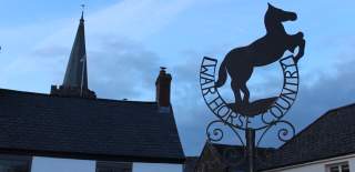 Silhouette of a horse sculpture with "War Horse Country" text, set against a twilight sky. Church spire and rooftops in the background convey a serene rural scene.