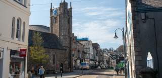 A quaint street scene features a stone church with a tall clock tower, flanked by shops and buildings; people walk under a bright blue sky.