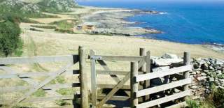 A wooden gate opens to a coastal path with distant green hills and a vast, calm blue sea. The scene conveys a serene, inviting atmosphere.§