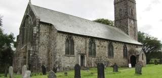 A historic stone church with tall arched windows and a square bell tower stands amidst a green graveyard. The scene feels tranquil under a cloudy sky.
