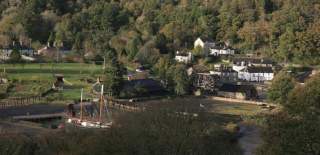 A scenic village nestled in a lush, forested valley. Features quaint cottages, a waterwheel, open green fields, and a docked ship, evoking tranquillity.