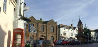A picturesque street scene features a red telephone box, stone buildings, parked cars, and a clock tower with a spire under a clear blue sky.