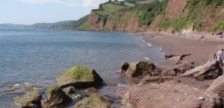 Rocky coastline with red cliffs and green hills, overlooking a calm blue sea. People stroll along the sandy shore under a bright, clear sky.