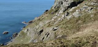Rocky cliffside overlooking a vast, calm blue ocean under a clear sky. The foreground features rugged, grassy terrain, creating a serene, natural scene.