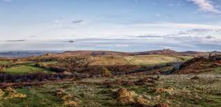 Expansive landscape with rolling hills, grassy fields, and patches of brown heather under a clear blue sky. The scene conveys a tranquil, open feel.