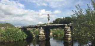 A person walks across a rustic stone bridge over a calm river, with lush green foliage and a partly cloudy blue sky, conveying a serene, peaceful scene.