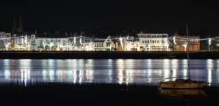 Nighttime waterfront scene in Bideford, North Devon, with buildings adorned with festive lights and their reflections on the water. A lone sailboat is silhouetted in the foreground, creating a serene atmosphere.
