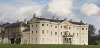 A grand historic Saltram House, cream-colored walls and tall windows set against a blue sky with fluffy clouds, surrounded by green lawns and leafless trees.