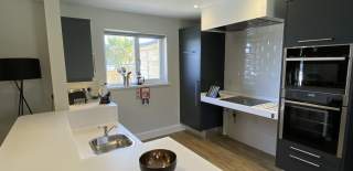 Modern kitchen with navy cabinets, white island, and three white stools. Wood flooring, black lamp, wall shelves, and pendant lights add a cosy feel