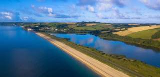 A serene coastal landscape with a long, narrow beach separating calm blue waters and lush green fields under a partly cloudy sky.