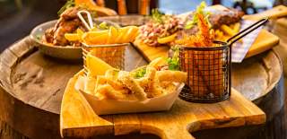A rustic table holds a wooden board with crispy fries in a metal basket, calamari, and ribs with garnish. The scene is warm, cosy, and inviting.