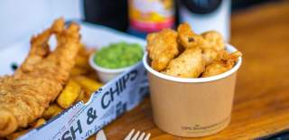 A hearty serving of fish and chips with mushy peas in a newspaper-lined box on a wooden table. A paper cup with fried snacks is nearby, evoking a warm, casual dining experience.
