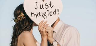 Newlyweds hold a heart-shaped sign reading "just married!" obscuring their faces. The bride wears a flower crown, exuding joy against a clear sky.