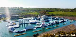 Inflatable water obstacle course on a calm lake, surrounded by lush greenery and bright sunlight. The scene feels adventurous and lively.
