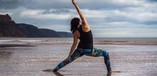 Woman practising yoga on a serene, sandy beach under a cloudy sky. She is in a warrior pose, wearing colourful leggings, conveying peace and focus.
