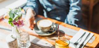 A close up of a person sat at a table with a coffee cup in front of them. The sun is casting light across the table and there is also a small vase with flowers.