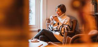 A woman sat on a comfy chair, next to a window, sipping a coffee.