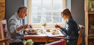 A man and a woman seated at a table opposite each other enjoying breakfast. There is a large window behind them and the sun is streaming in.