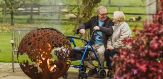 2 people sat on a bench behind a log burner  1 person holding a walking frame.