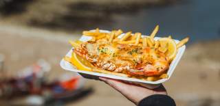 A hand holding a plate of lobster and fries overlooking Flamborough North Landing