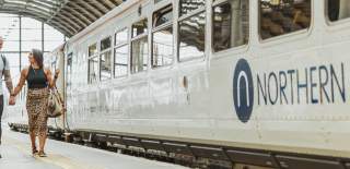 A couple walking hand-in-hand along a train station platform. A Northern train is stationary on the tracks beside them.