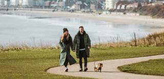 A couple walking hand-in-hand along a path on the Sewerby cliff tops in Bridlington. Their small sausage dog is walking beside them on a lead and you can see a view of Bridlington beach in the background.