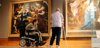 A woman in a wheelchair and a man using crutches viewing two large paintings inside Ferens Art Gallery. The spacious gallery features well-lit artwork and an accessible environment.