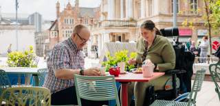 A woman in a wheelchair and a man sitting outside Ponto Lounge in Queen Victoria Square, Hull, enjoying a sunny day in a vibrant public space.