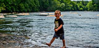 A young boy playfully stands in the Chattahoochee River water, smiling brightly under the warm sun.