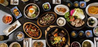 A colorful spread of Korean dishes including bibimbap, grilled meat, kimchi, and various side dishes arranged on a dark table.