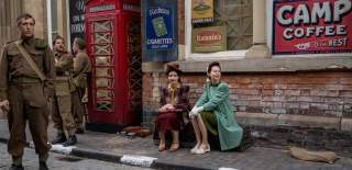 Actors dressed in 1940s clothing sit beside a vintage British red telephone box on a cobbled street, surrounded by period military uniforms and retro advertising signs.