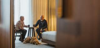 Hotel room scene with two guests seated at a small table enjoying drinks, while a dog lies on the carpeted floor beside the bed, with warm lighting and floor‑to‑ceiling curtains creating a relaxed atmosphere.